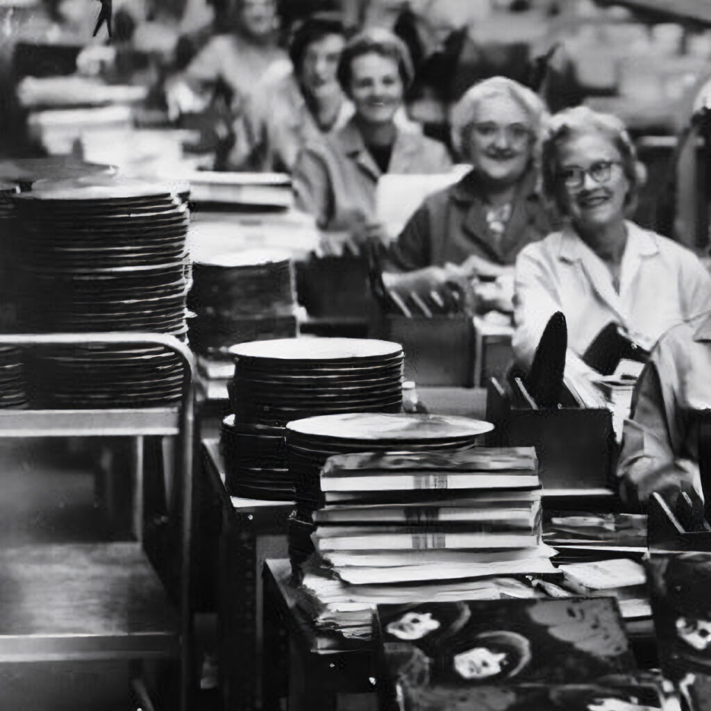 Women inspecting and stacking vinyl records on an assembly line at a record pressing plant in the mid-20th century.