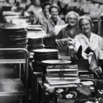 Women inspecting and stacking vinyl records on an assembly line at a record pressing plant in the mid-20th century.
